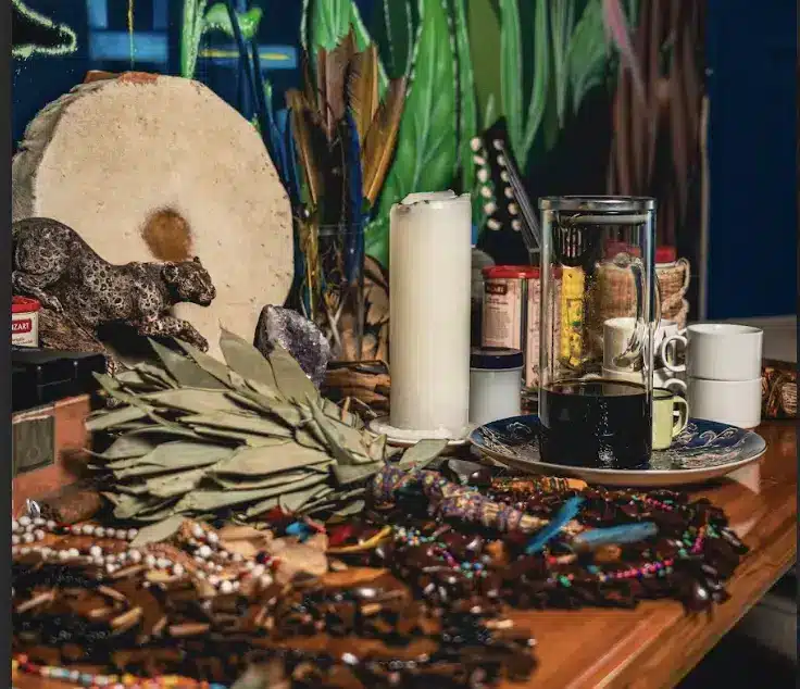 ceremonial altar with the candle, ayahuasca medicine, cups, cascavelles, the waira and more atop the altar. Artwork of the jungle in the background.