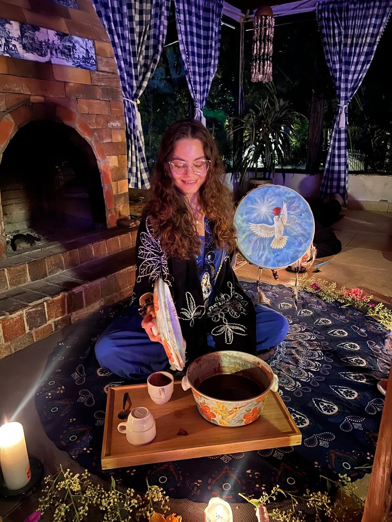 Facilitator seated by a brick fireplace at an evening cacao ceremony in Medellin, a bowl of ceremonial cacao and cups on a tray beside her and a painted drum behind