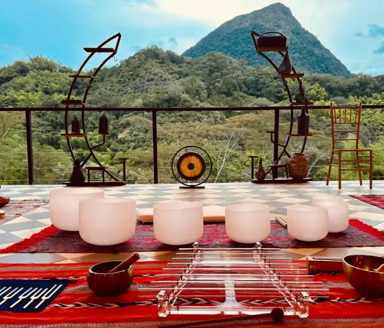 A sound bath set up on a terrace facing Cerro Tusa: crystal bowls, a gong, tuning forks and a glass harp on red rugs, with the pyramid mountain and green valley behind